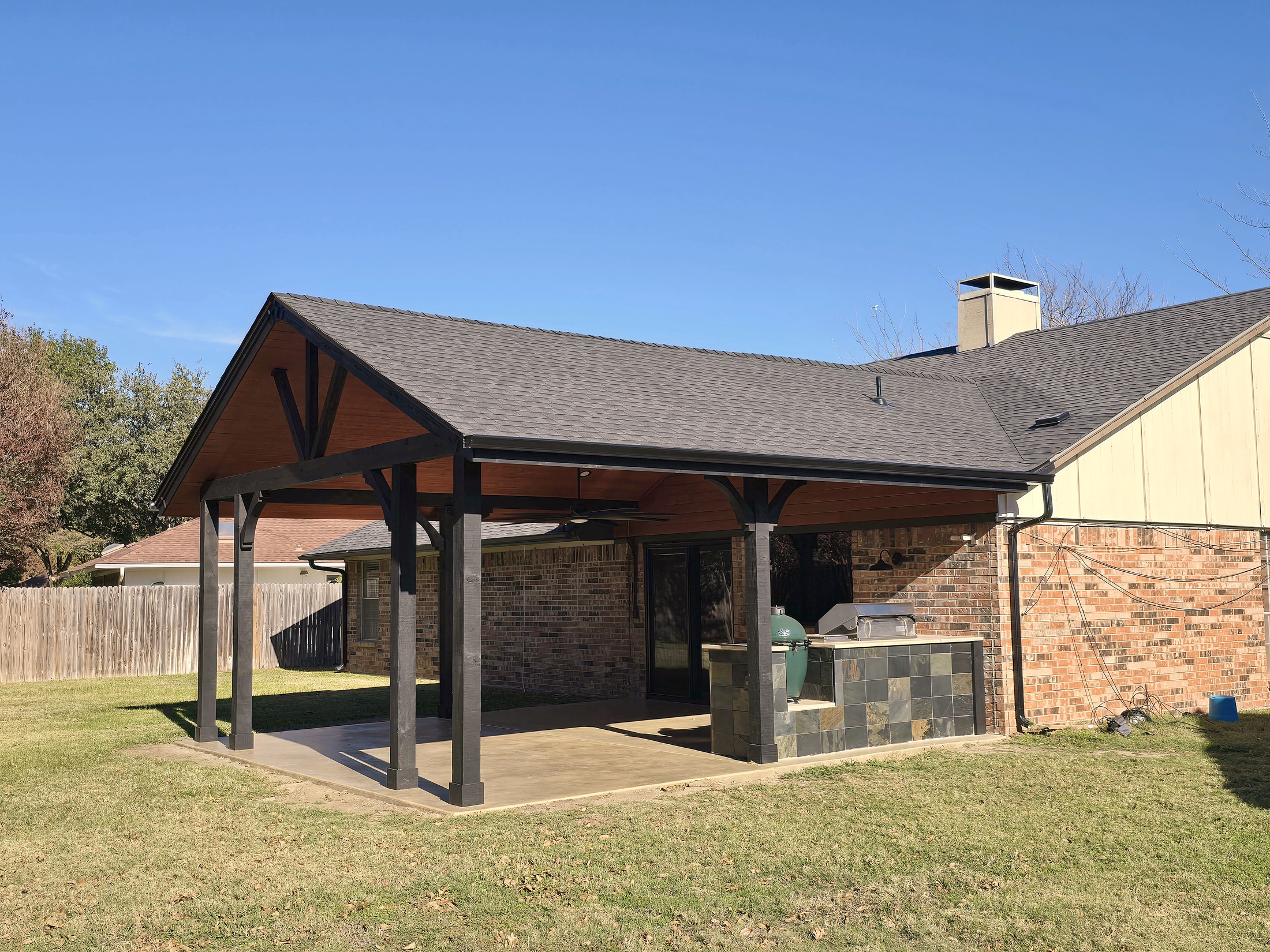 A cedar patio cover with a shingled gable roof on the back of a brick home